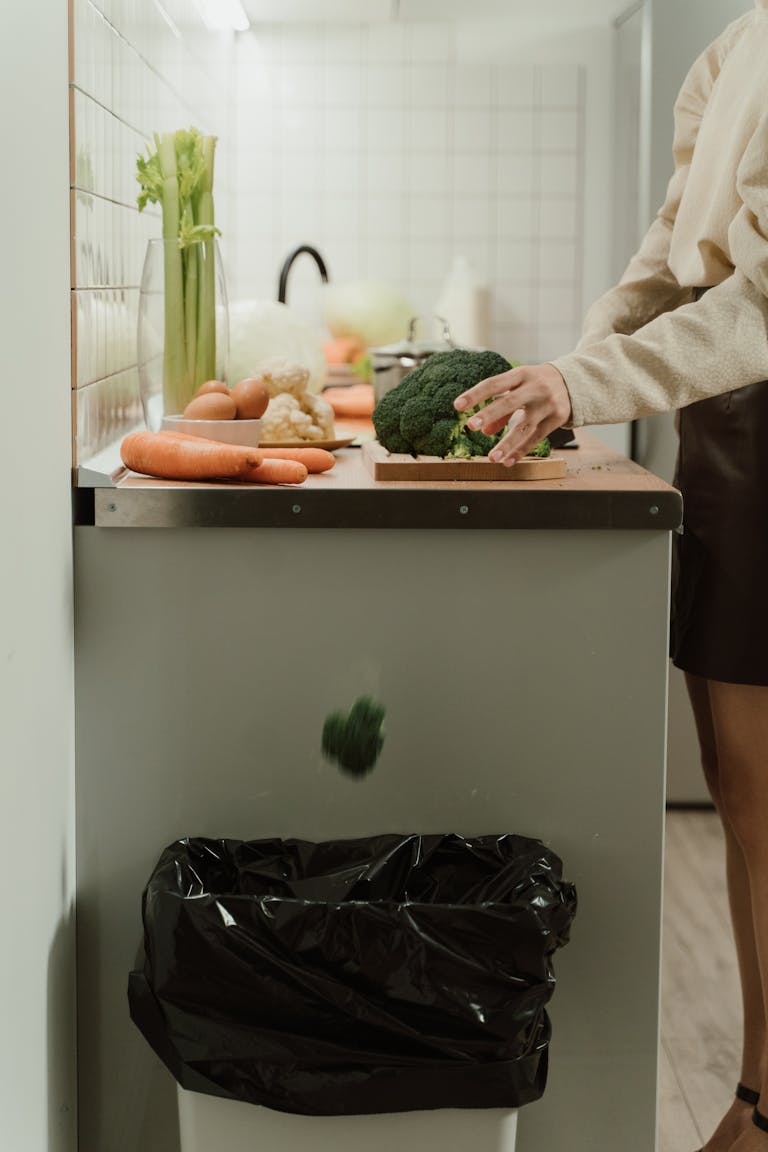 Person discarding broccoli in a stylish kitchen with fresh vegetables on the counter.